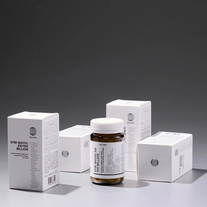 Eine braune Pillenflasche mit der Aufschrift Sym-Biotic 23/100 Billion ist von vier weißen Produktschachteln mit ähnlicher Aufschrift umgeben, die alle auf einer grauen Fläche vor einem grauen Hintergrund stehen.