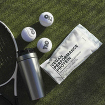 Eine Shakerflasche, ein Tennisschläger, drei Tennisbälle und eine Packung DALUMA VEGANES PERFORMANCE PROTEIN KAKAO liegen auf grünem Kunstrasen in hellem Sonnenlicht.