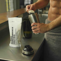 Nach dem Training gibt eine mit einem Handtuch bekleidete Person veganes Proteinpulver in den DALUMA PROTEIN SHAKER EDELSTAHL auf der Küchentheke und entscheidet sich für diese nachhaltige Alternative mit einem Performance Protein Beutel in der Nähe.
