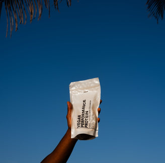 Eine Hand hält eine weiße DALUMA VEGANES PERFORMANCE PROTEIN NEUTRAL-Tüte vor einem klaren blauen Himmel hoch, in der linken oberen Ecke sind Palmenblätter zu sehen.