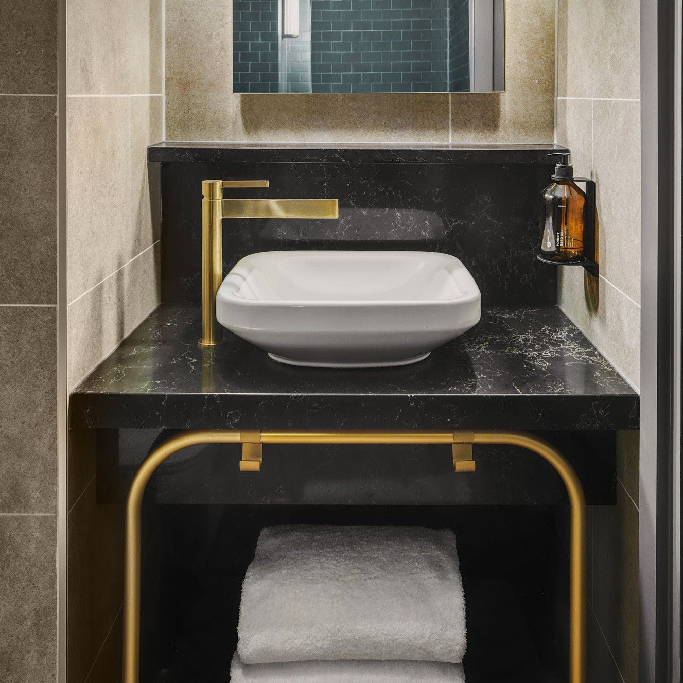 Die DALUMA BODYCARE WANDHALTERUNG bietet ein modernes Design mit einer Arbeitsplatte aus schwarzem Marmor, einem weißen, rechteckigen Waschbecken, einem goldenen Wasserhahn, einem wandmontierten Seifenspender und ordentlich gefalteten weißen Handtüchern auf einer goldgerahmten Ablage darunter.