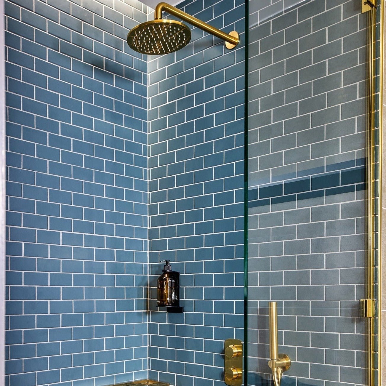 Eine moderne Dusche mit blauen Kacheln, einer goldenen Regendusche, einer Glaswand und der DALUMA BODYCARE WANDHALTERUNG - einem kleinen schwarzen Regal mit einer dunklen Flasche.
