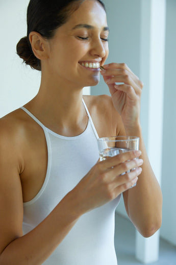 Frau lächelt, während sie eine Pille mit einem Glas Wasser einnimmt. Sie trägt ein weißes Tank-Top und steht in einer hellen Innenumgebung.